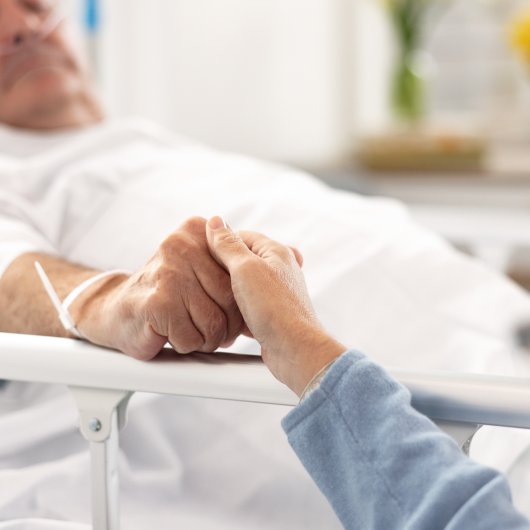 End of life man lying in a hospital bed and holding his partner's hand.