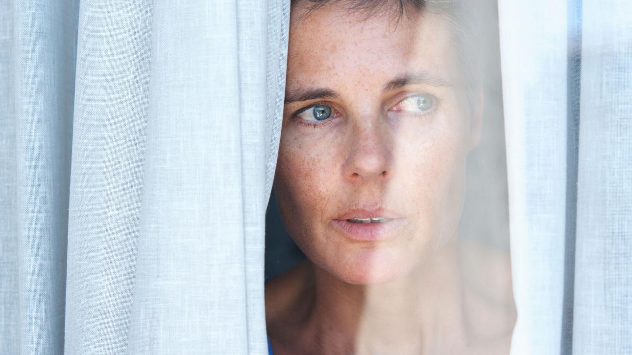 A worried woman looks between the curtains of a window