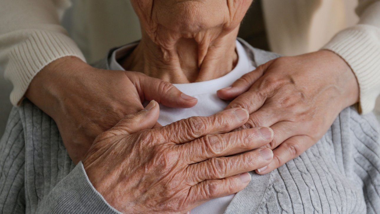 Dame âgée enlacée tendrement par un proche se tenant derrière elle