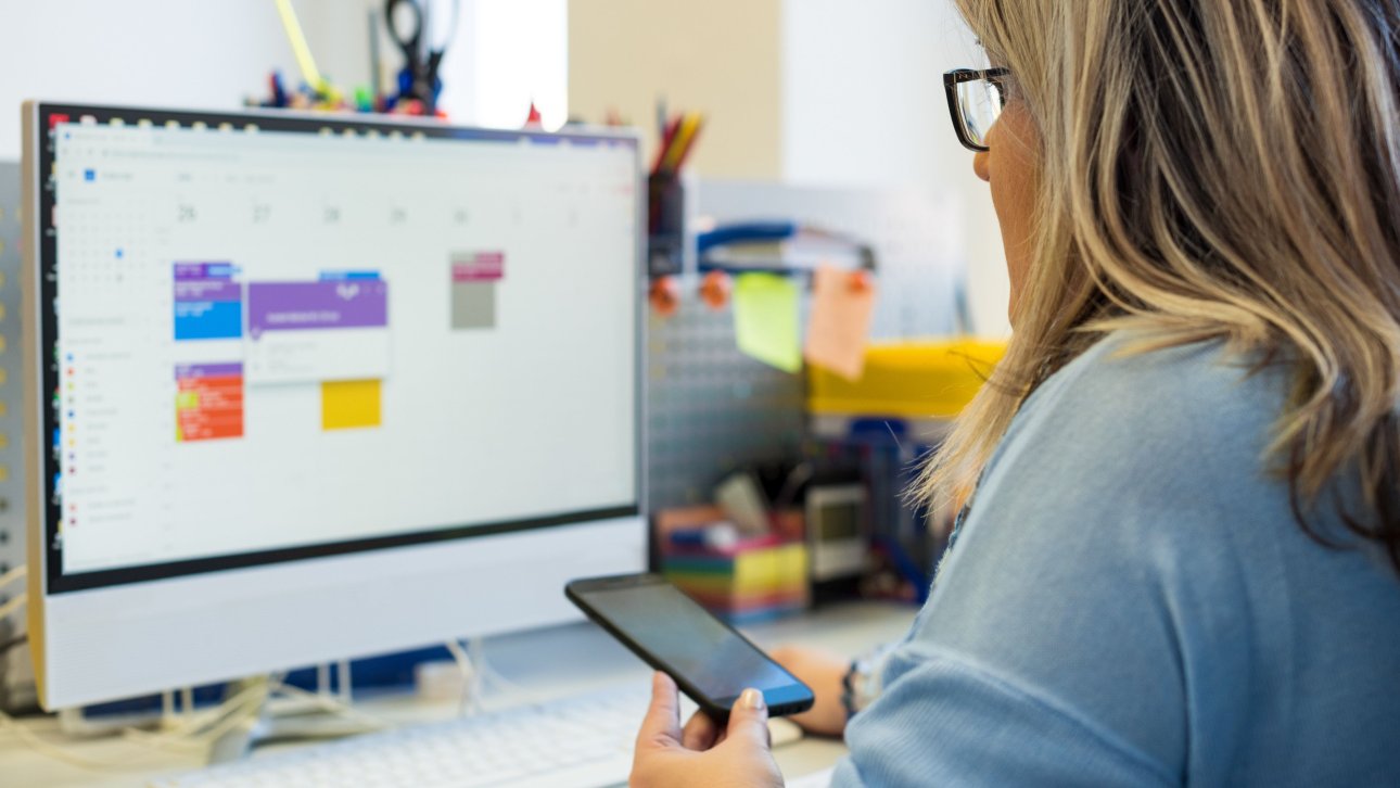 Femme tenant son cellulaire en regardant un calendrier affiché sur son ordinateur