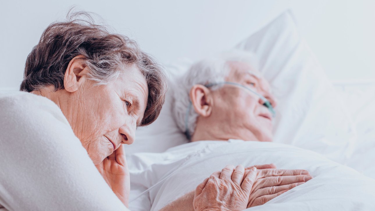 Elderly lady displaying a sad expression at the bedside of her sick and bedridden spouse.