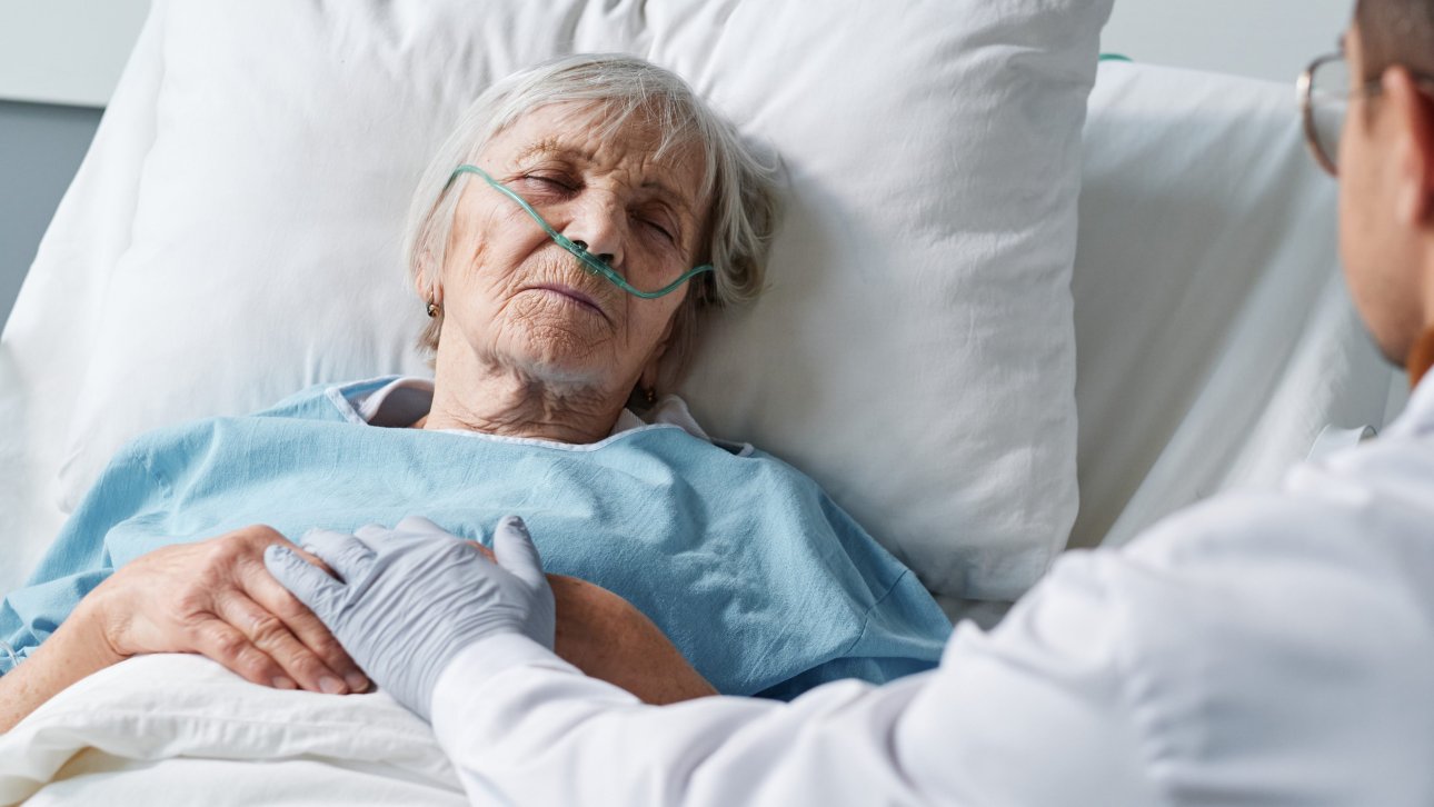 Dame âgée en fin de vie allongée dans son lit d'hôpital et se faisant réconforter par le médecin.