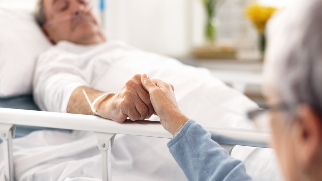 End of life man lying in a hospital bed and holding his partner's hand.