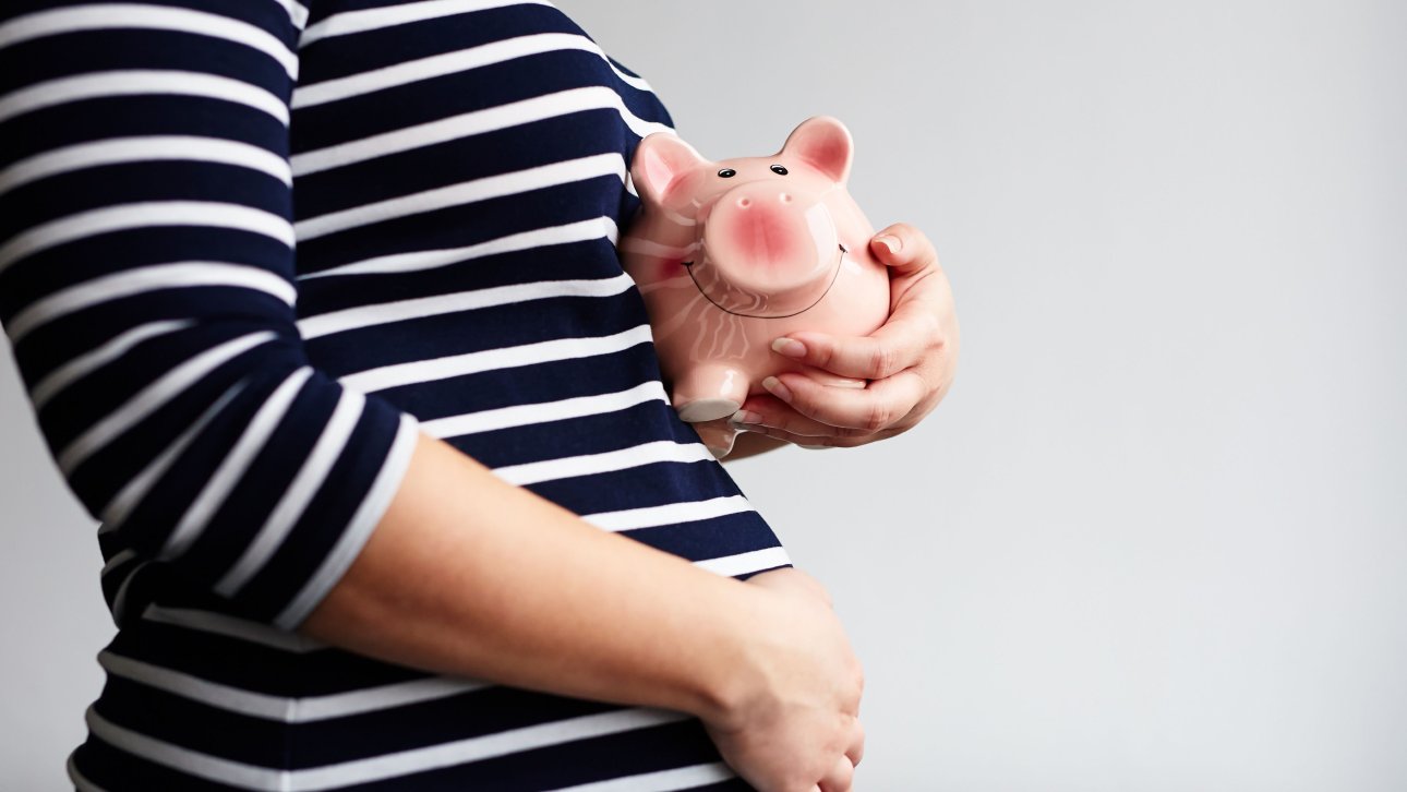 Pregnant woman touching her belly with one hand and holding a piggy bank with the other.