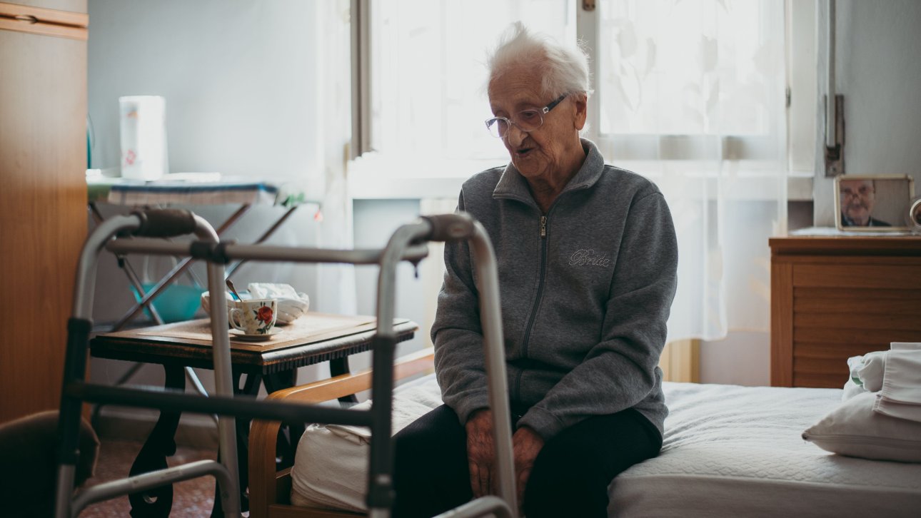 Elderly person sitting on the edge of a bed looking sadly at their walker.