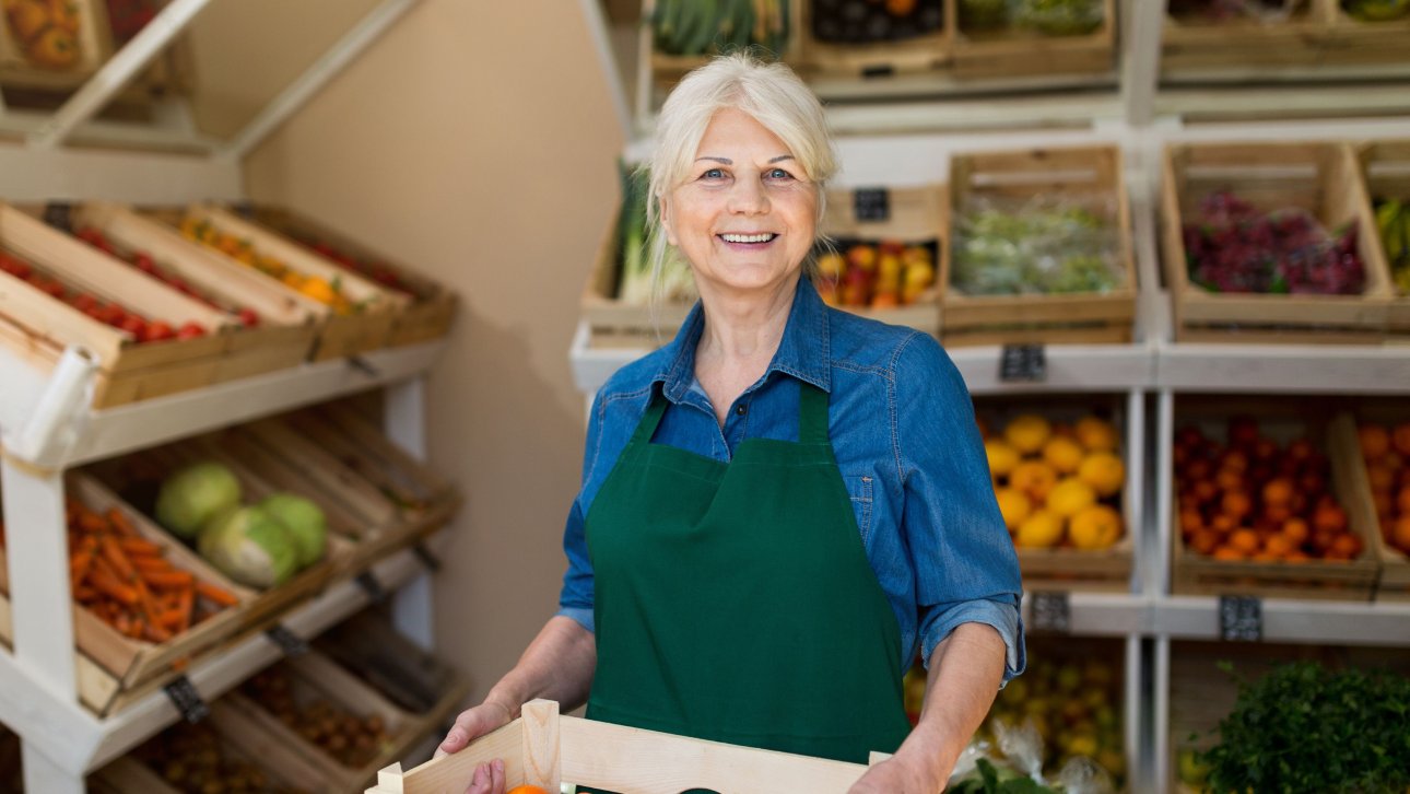 Smiling lady aged around 60 wearing an apron and holding a bin of vegetables in a grocery store.