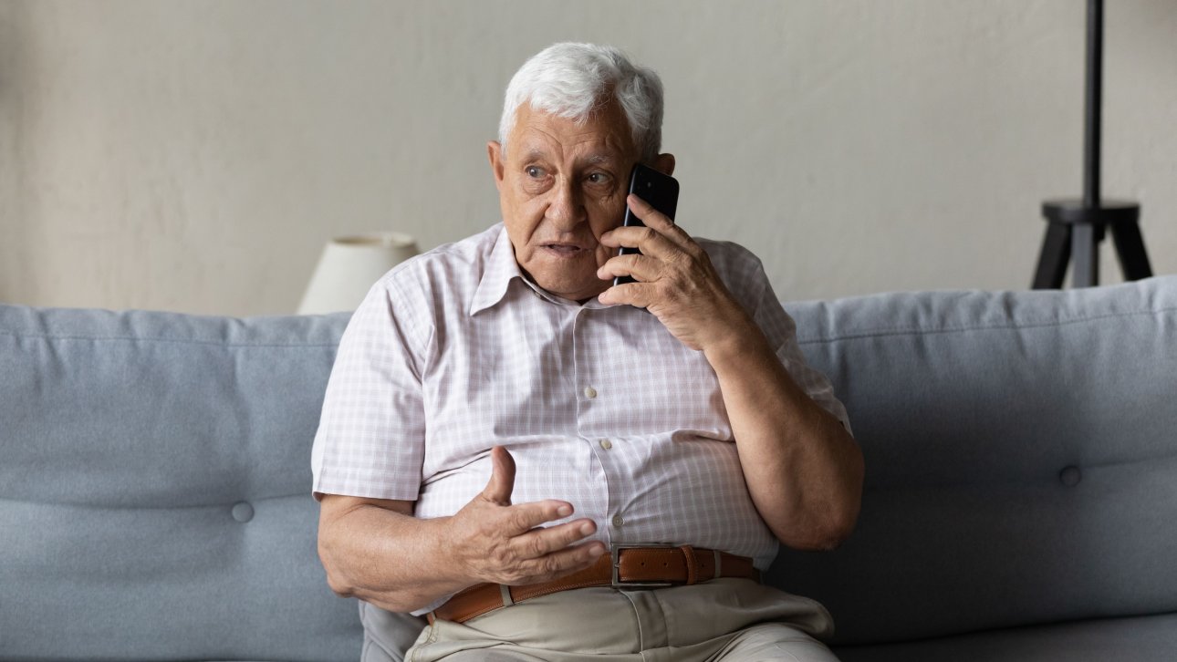 Elderly man sitting on his couch and chatting on the phone with a concerned look.