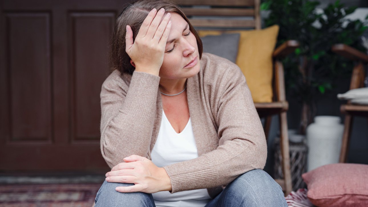 Discouraged looking woman sitting on her stoop with her hand on her forehead.