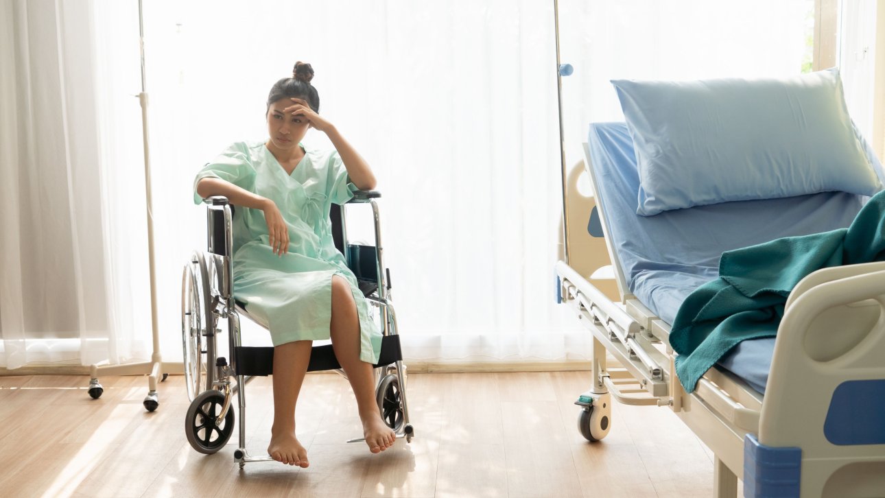 Upset young woman sitting in a wheelchair and wearing a hospital gown.