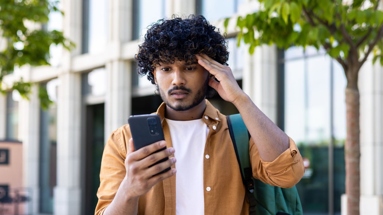 Un étudiant sortant de l'école regarde son cellulaire d'un air surpris et angoissé.