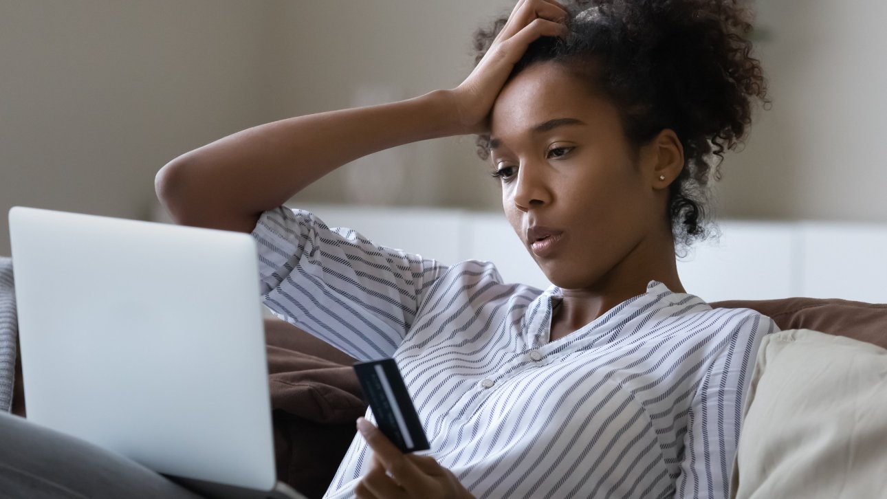 Jeune femme tenant sa carte de crédit en regardant son ordinateur portable d'un air confus.