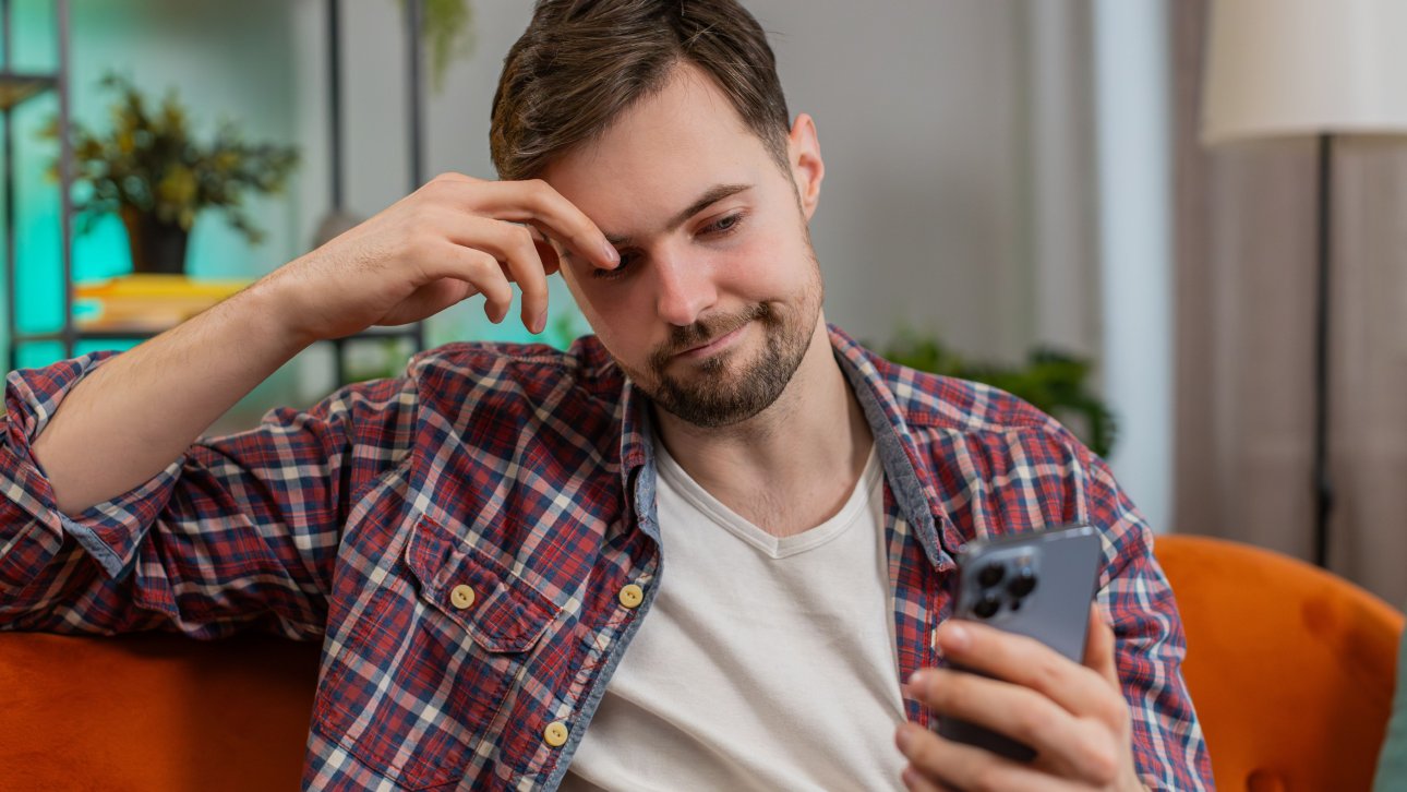 A man sitting in an armchair looks annoyed as he stares at his mobile phone screen.