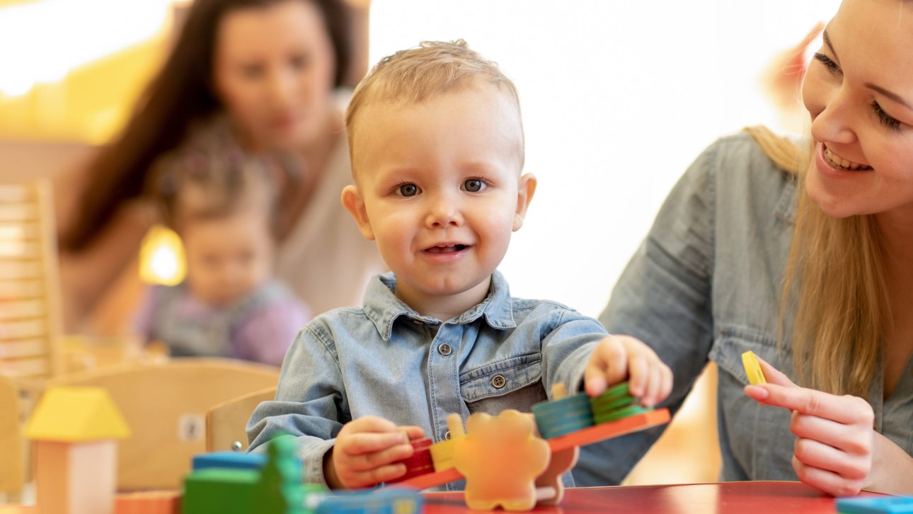 Petit garçon assis devant des jouets aux côtés de son éducatrice à la garderie ou au CPE.