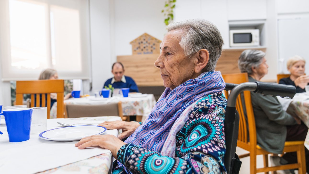 Une dame âgée est assise devant une assiette vide dans un centre d'hébergement pour aînés.