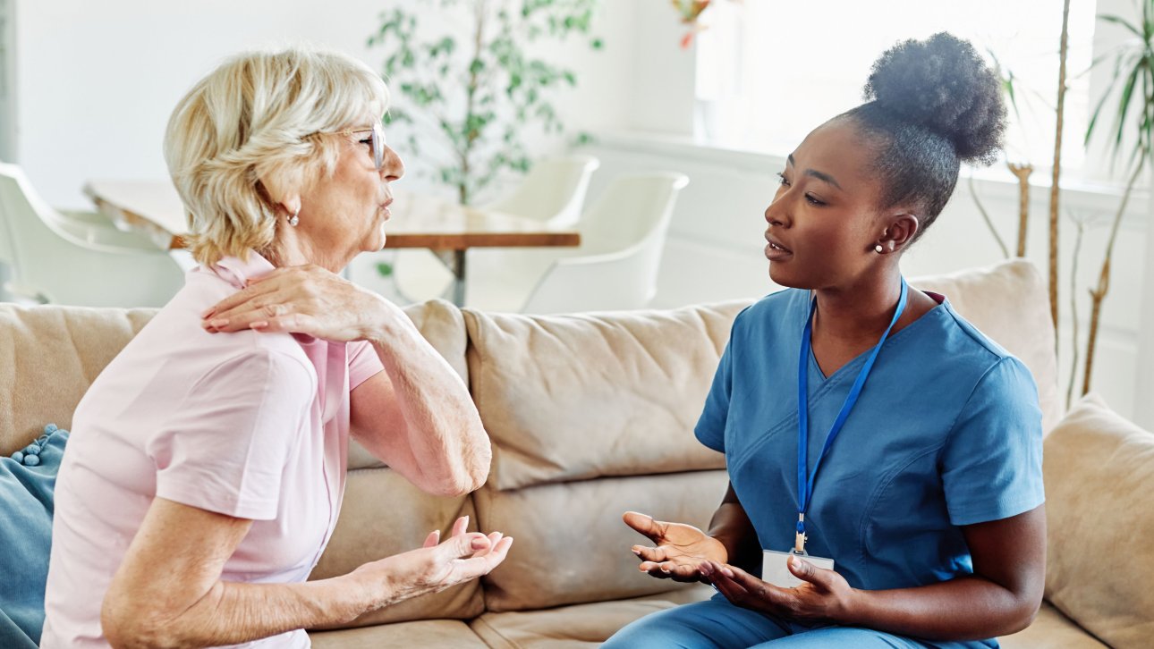Dame exprimant de la douleur en montrant son épaule à l'infirmière qui lui offre des soins à domicile.