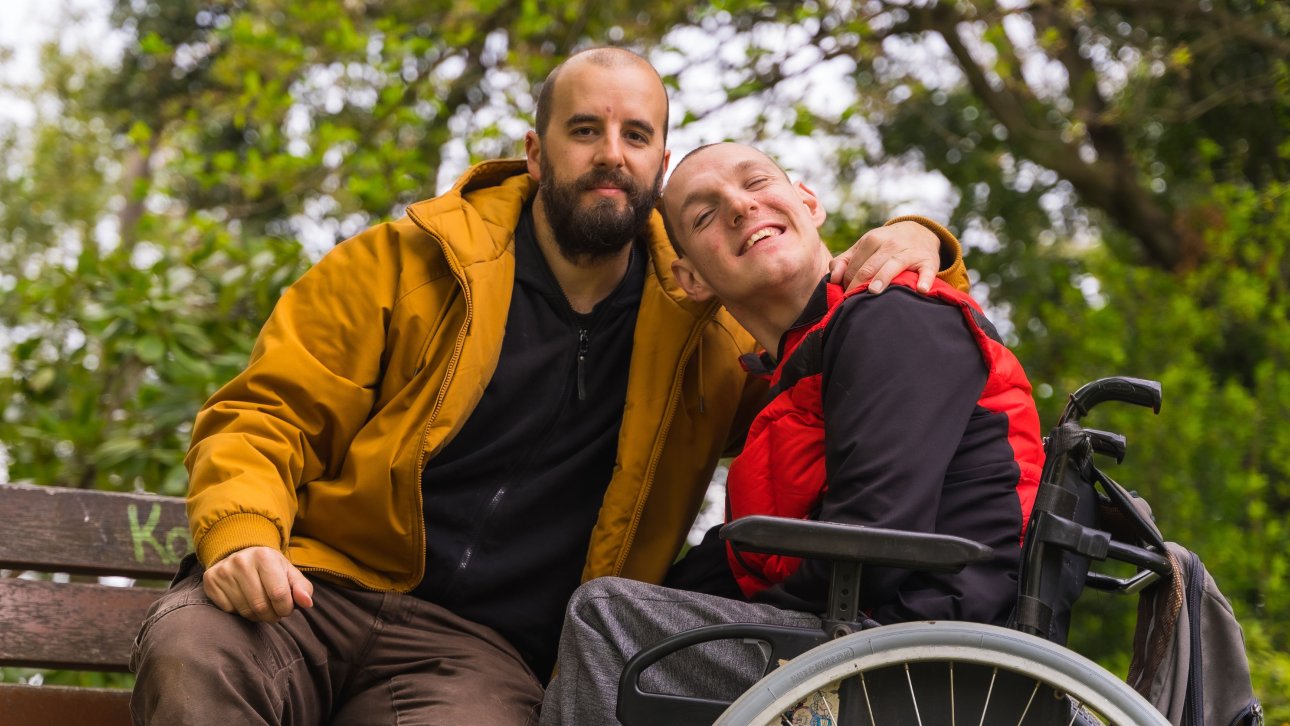 Un père entoure de son bras son fils en fauteuil roulant en souriant à la caméra.
