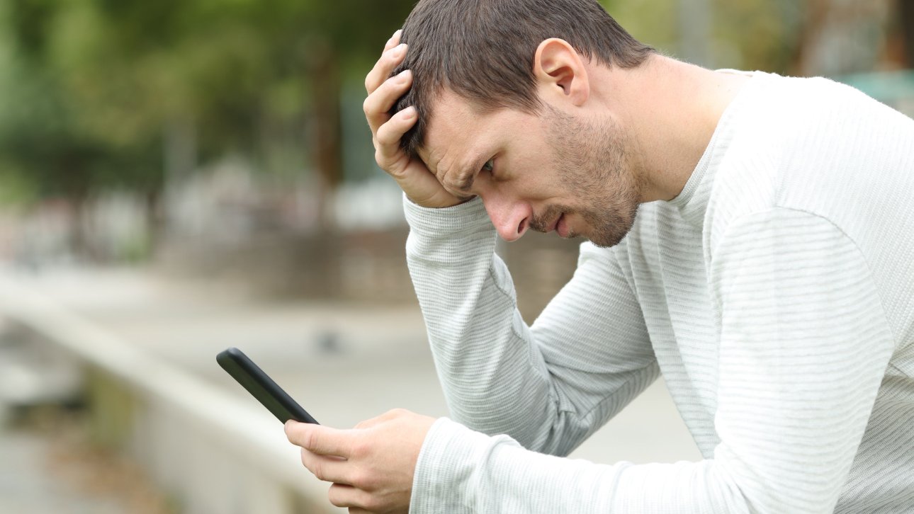 A man sitting on a bench outside is looking at his mobile phone screen, looking worried