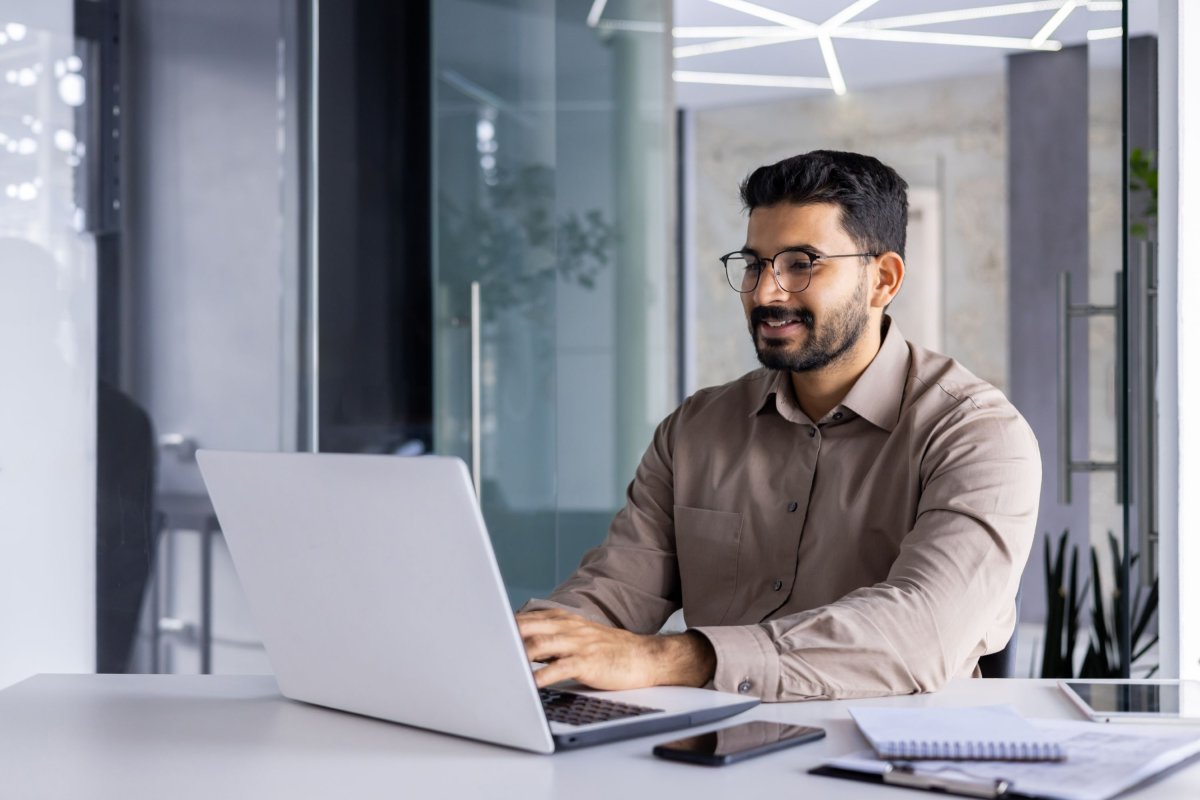 Employé souriant travaillant sur un ordinateur portable placé près de plusieurs documents.