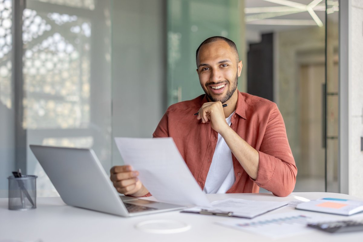 Un homme souriant travaille devant son ordinateur en tenant un document d'une main.