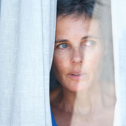 A worried woman looks between the curtains of a window