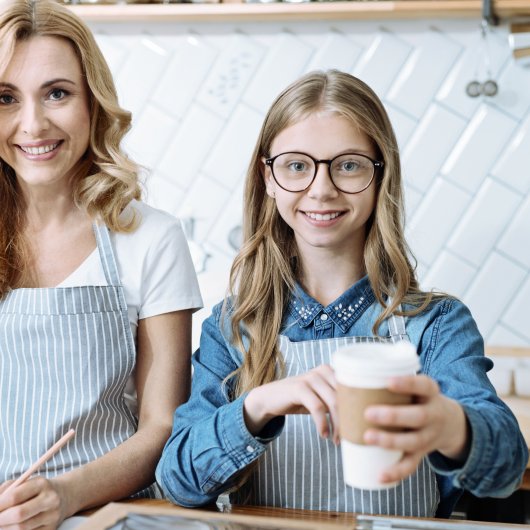 Mother working behind a counter alongside her approximately 14 year old daughter serving coffee to a customer