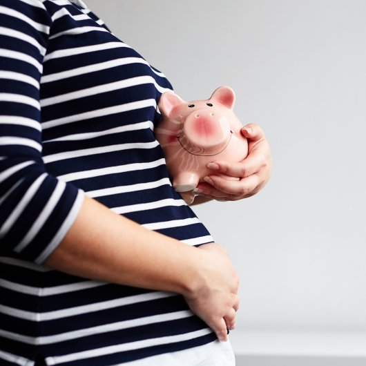 Pregnant woman touching her belly with one hand and holding a piggy bank with the other.