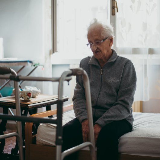 Elderly person sitting on the edge of a bed looking sadly at their walker.