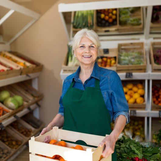 Smiling lady aged around 60 wearing an apron and holding a bin of vegetables in a grocery store.