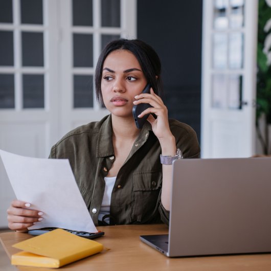 Femme contrariée assise devant son ordinateur et parlant au cellulaire en tenant un document.