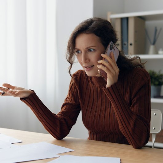 A woman sitting at a table covered with documents is having a serious conversation on the phone.