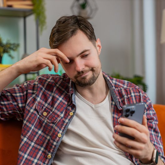 A man sitting in an armchair looks annoyed as he stares at his mobile phone screen.