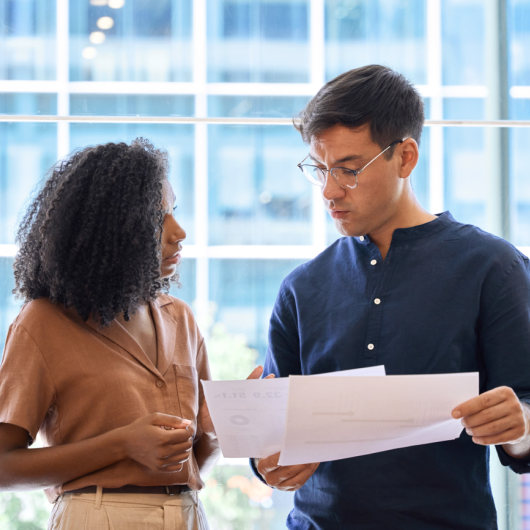 Two colleagues are standing side by side and chatting while looking at documents.