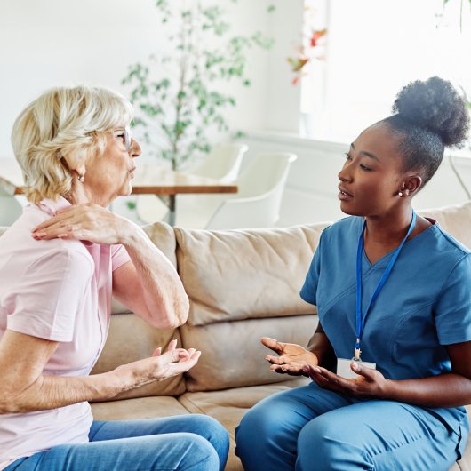 Dame exprimant de la douleur en montrant son épaule à l'infirmière qui lui offre des soins à domicile.
