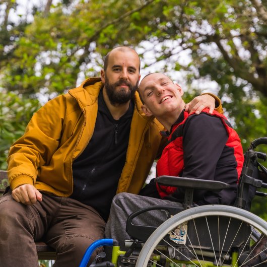 Un père entoure de son bras son fils en fauteuil roulant en souriant à la caméra.
