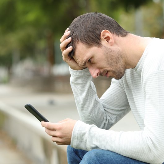 Un homme assis sur un banc à l'extérieur consulte l'écran de son cellulaire, l'air inquiet